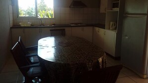 A kitchen with a round dining table, chairs, a refrigerator, and a window with a view of greenery. - Gîte Océania (Wallis Island)