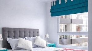 Un dormitorio con cabecera tapizada de color gris, colcha escocesa, mesitas de noche verde azulado y vistas a edificios a través de una ventana.