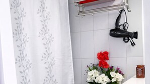 Un baño con inodoro, cortina de ducha, repisa con toalla roja, secador de pelo y florero.