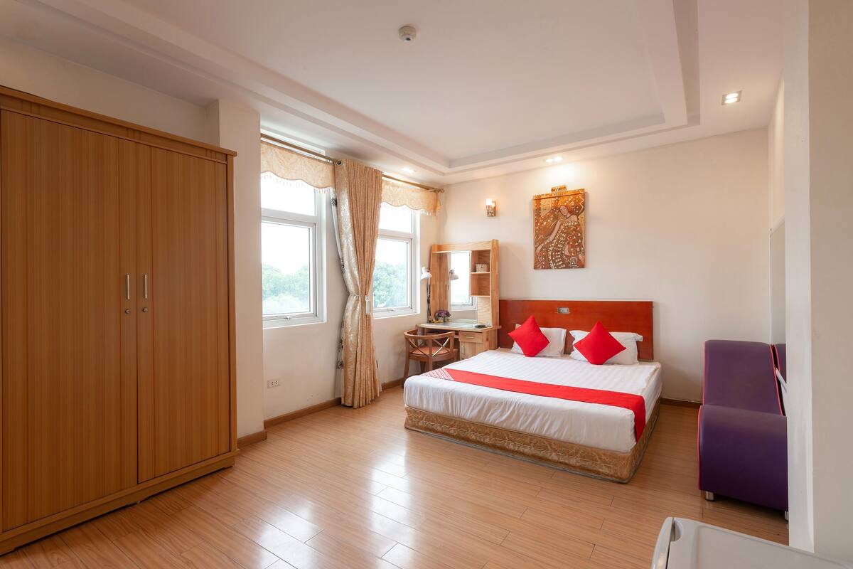 A hotel room with a large wardrobe, a bed with red pillows, a desk with a chair, and a window with curtains.