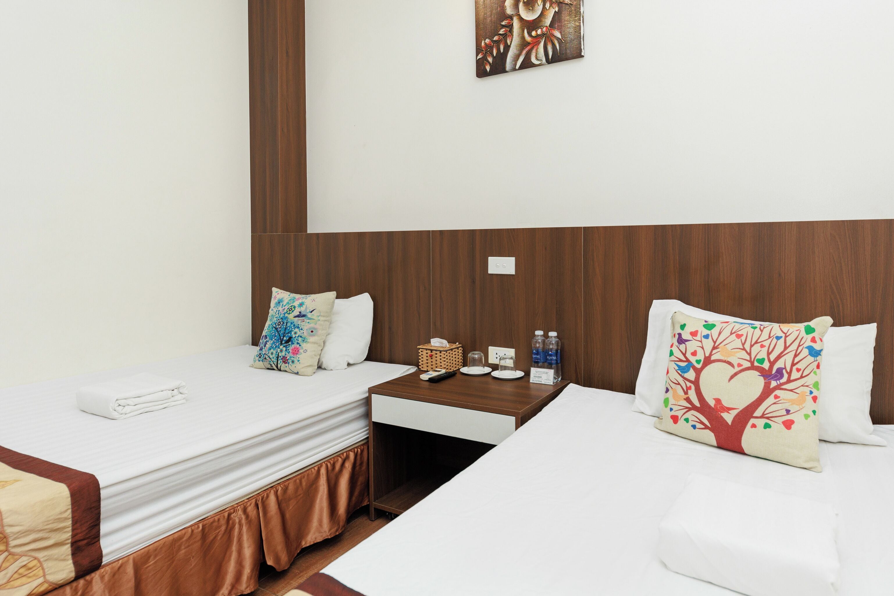 A hotel room with two single beds, a wooden headboard, a small bedside table with a basket and bottles, and a wall-mounted painting.
