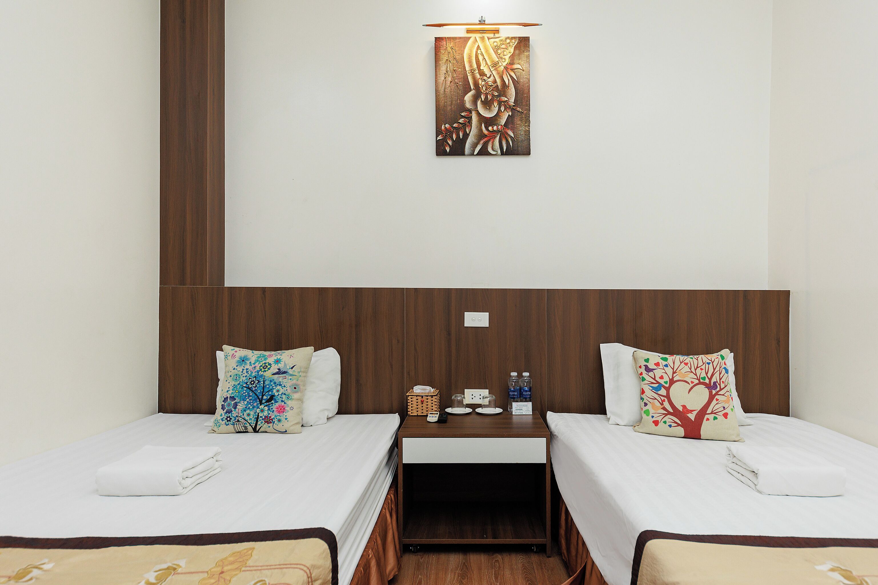 A hotel room with two single beds, a wooden headboard, a small table with a lamp, and a wall-mounted artwork.