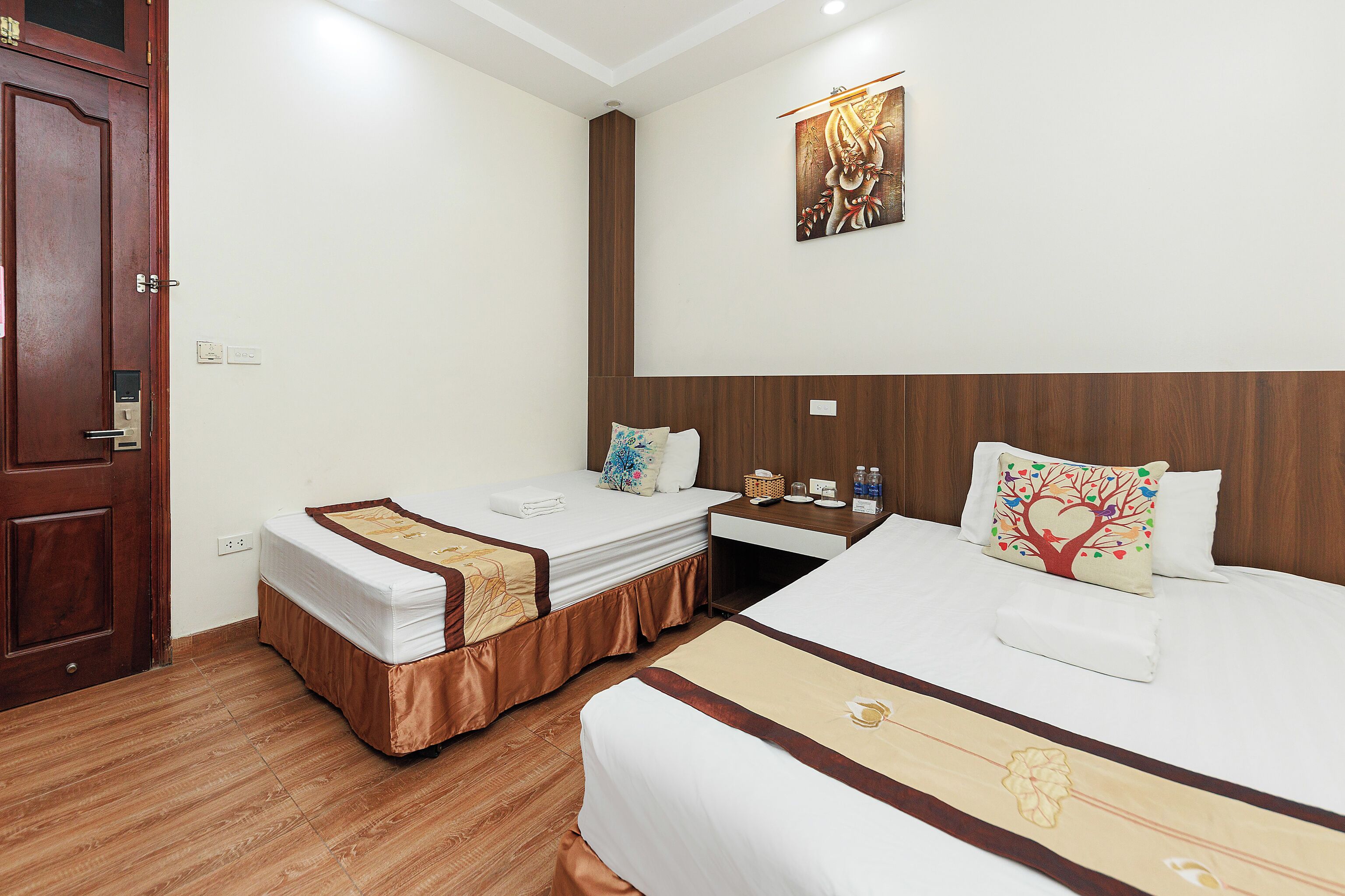 A hotel room with two single beds, a wooden door, a small desk with a chair, a wall-mounted artwork, and a wooden floor.