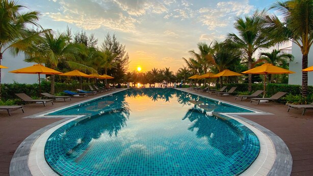 A swimming pool with a tiled surface, surrounded by lounge chairs and yellow umbrellas, with palm trees and a sunset in the background. - M Villas Phu Quoc (Phu Quoc)