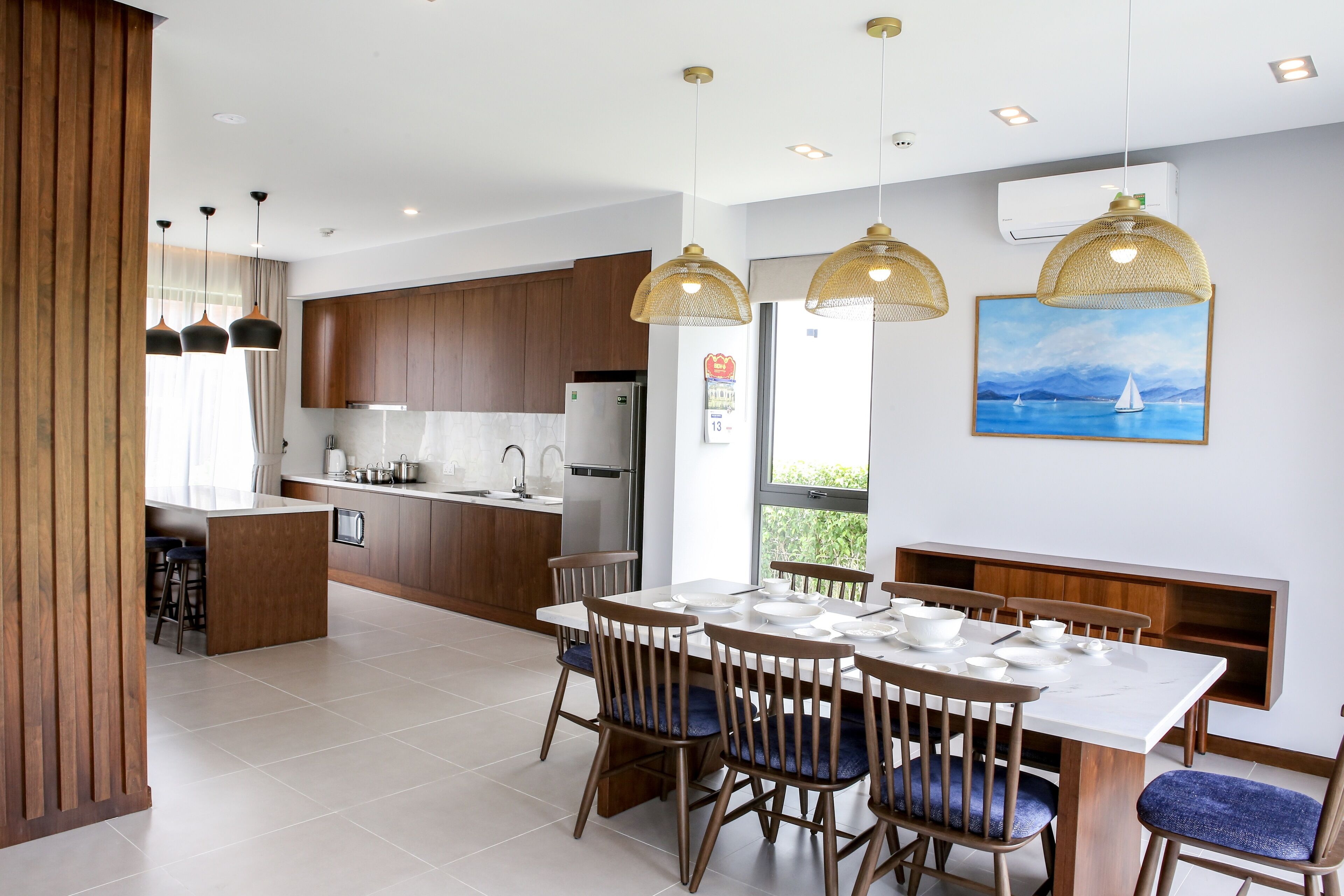A modern kitchen with wooden cabinets, a dining area with a white table and blue chairs, and pendant lights.