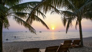 A beach with wooden lounge chairs and palm trees, overlooking the ocean at sunset. - M Villas Phu Quoc (Phu Quoc)