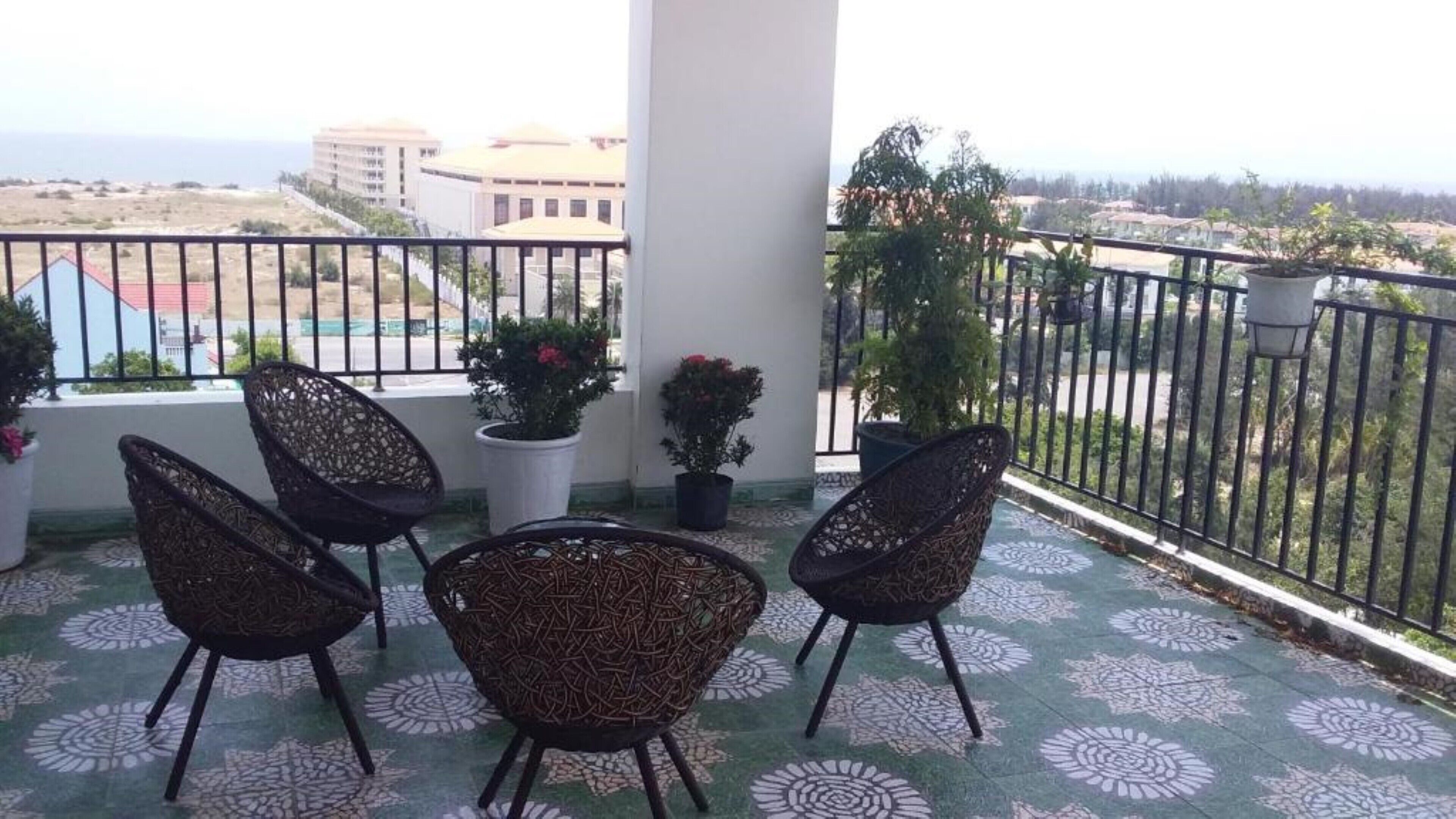 Un balcón con cuatro sillas de mimbre, plantas en macetas y vistas a edificios y al mar.
