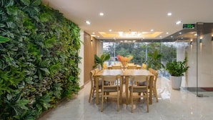 A dining area with a table and chairs, a green wall with plants, and a view through a glass door.