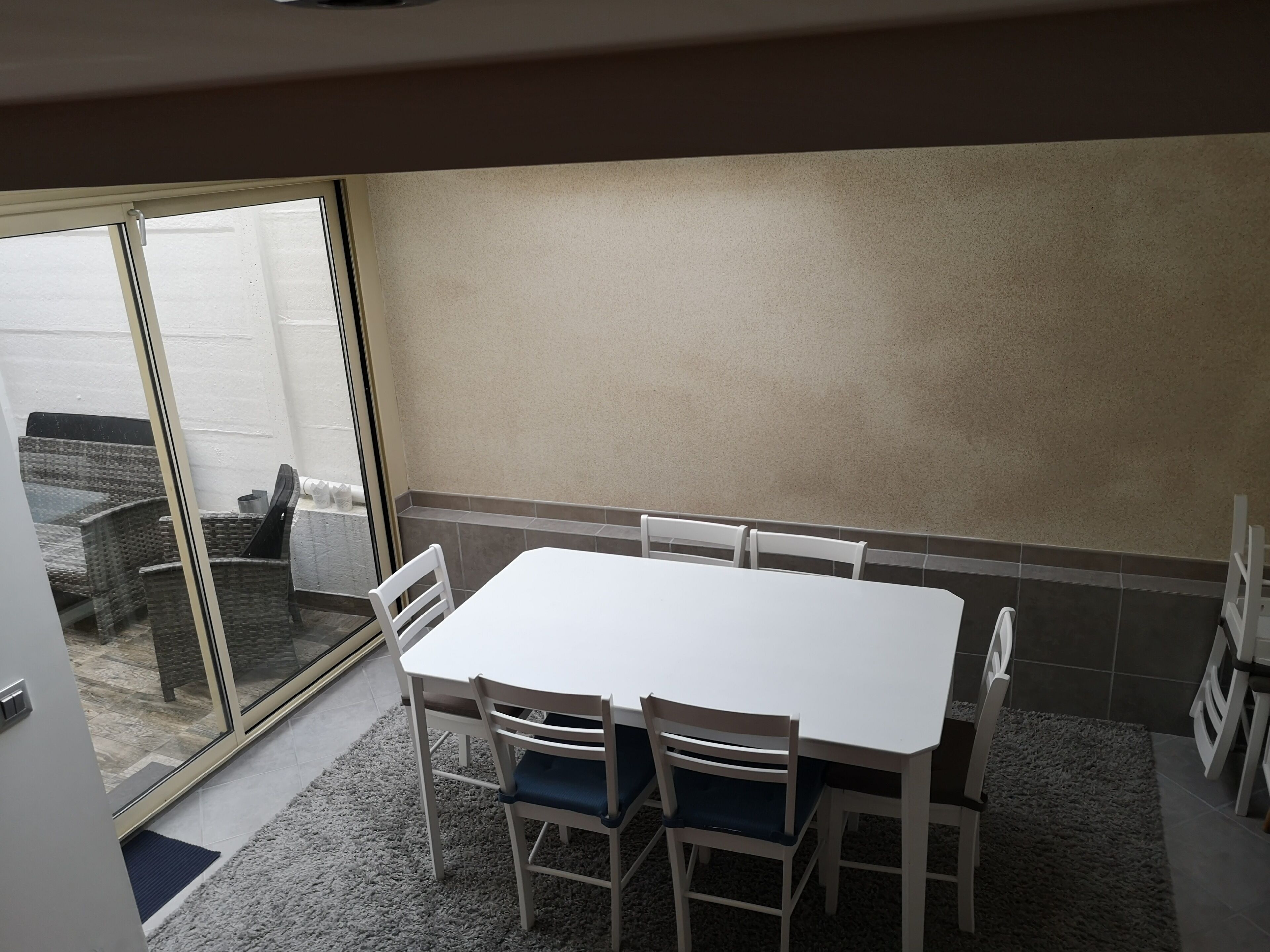 A dining area with a white table and chairs, a sliding glass door leading to a balcony, and a tiled wall.