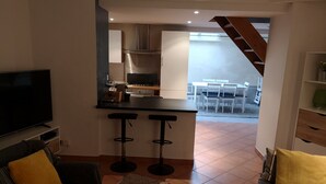 A modern living room with a TV, a sofa, a coffee table with a flower vase, and a kitchen area with a dining table and chairs. - GITE 7/8 P. LE NAPOLEON, Not suitable for PRMs (Saint-Leu-la-Forêt)