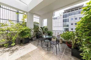 Een balkon met betegelde vloer, planten in potten, en een glazen tafel met metalen stoelen.