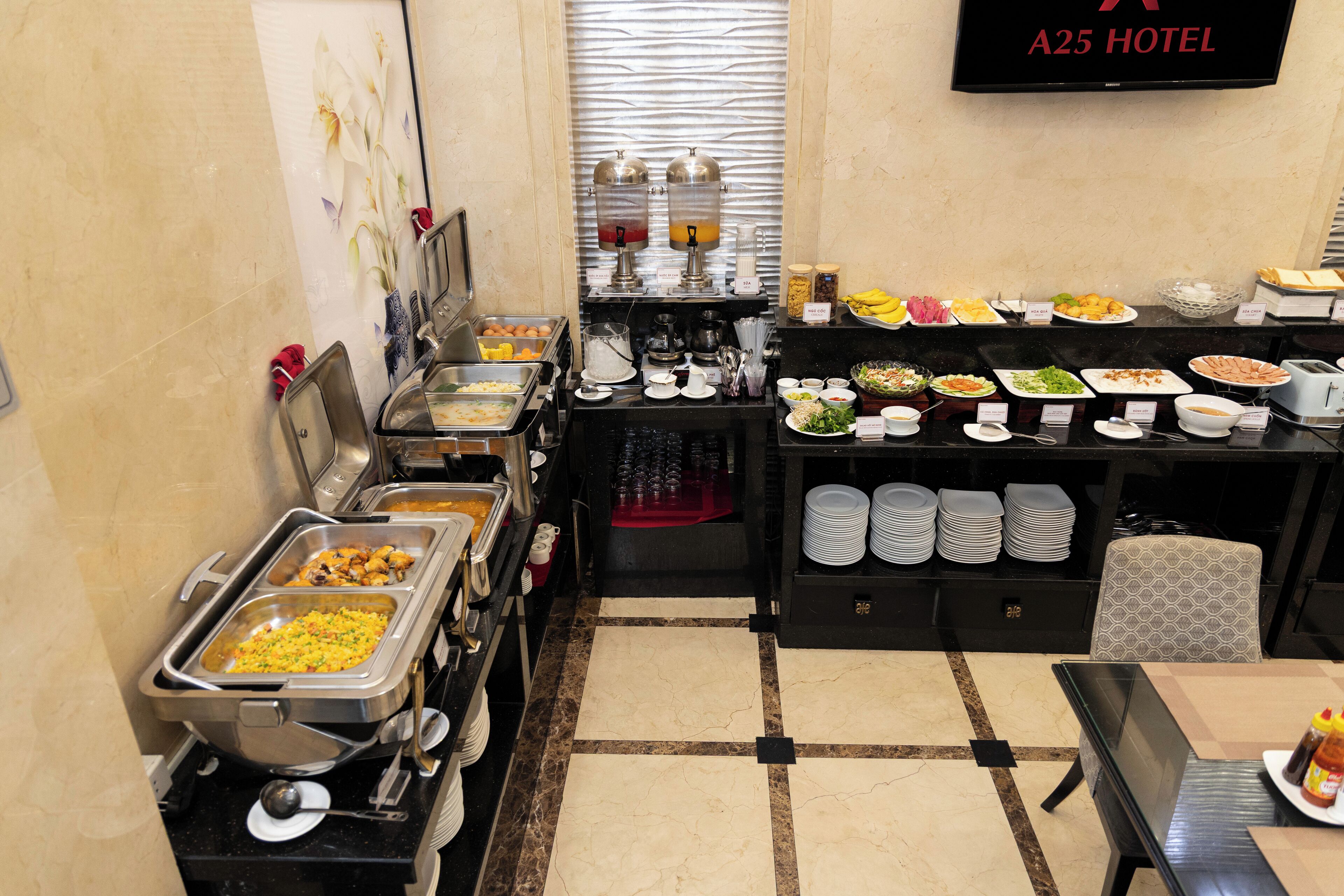 Ein Buffet mit verschiedenen Speiseauslagen, einem Getränkeautomaten und einem Fernseher, der den Namen des Hotels anzeigt.