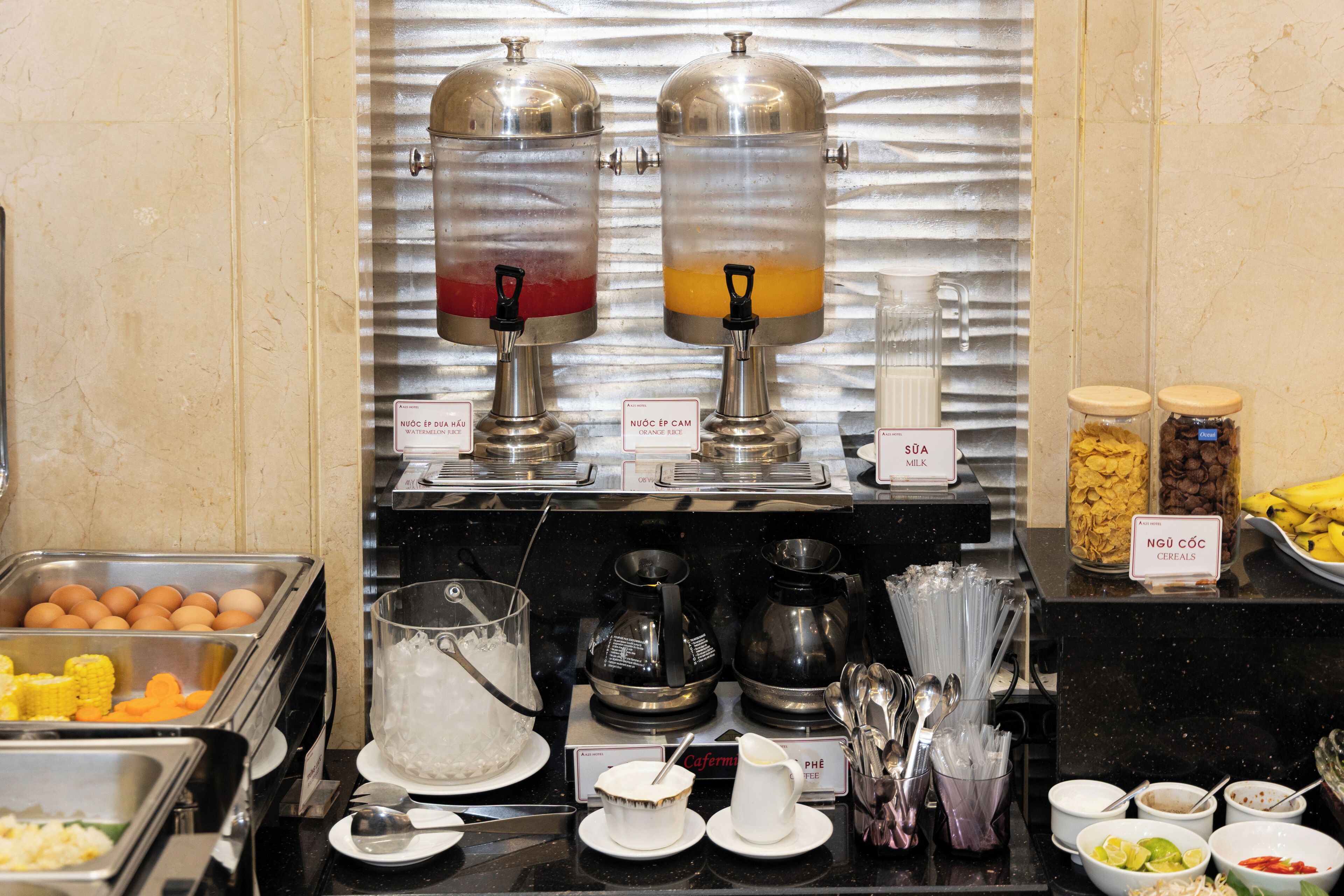 Un buffet proposant une variété de plats et de boissons, avec notamment un distributeur de jus de pastèque et d’orange, ainsi qu’un espace dédié aux céréales et au lait.