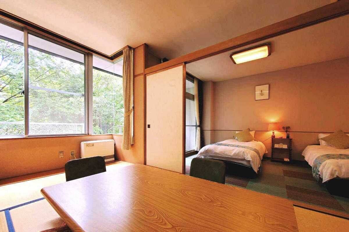 Een kamer met een houten tafel en stoelen, twee enkele bedden, een klein nachtkastje met een lamp, en een raam met uitzicht op groen.