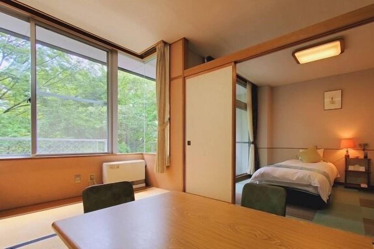 Een hotelkamer met een houten tafel, twee bedden, een raam met uitzicht op bomen en een plafondlamp.