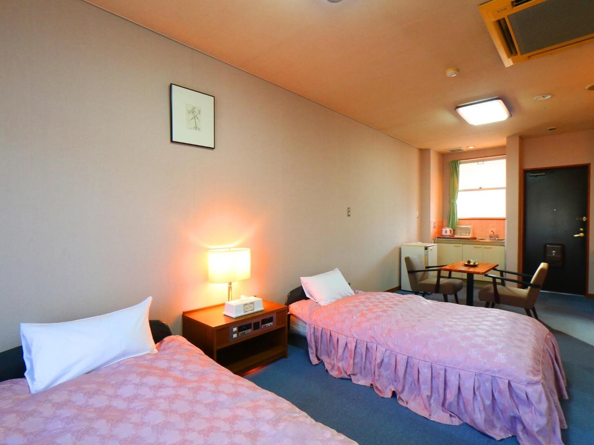 A hotel room with two beds, a desk, a chair, a small kitchenette, and a window with curtains.