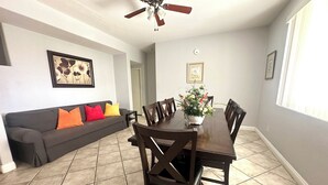 A dining area with a dark wooden table and chairs, a gray sofa with colorful pillows, a ceiling fan, and a potted plant on the table. - County Licensed STR! Comfortable, spacious home 15 minutes to The Strip (Las Vegas)