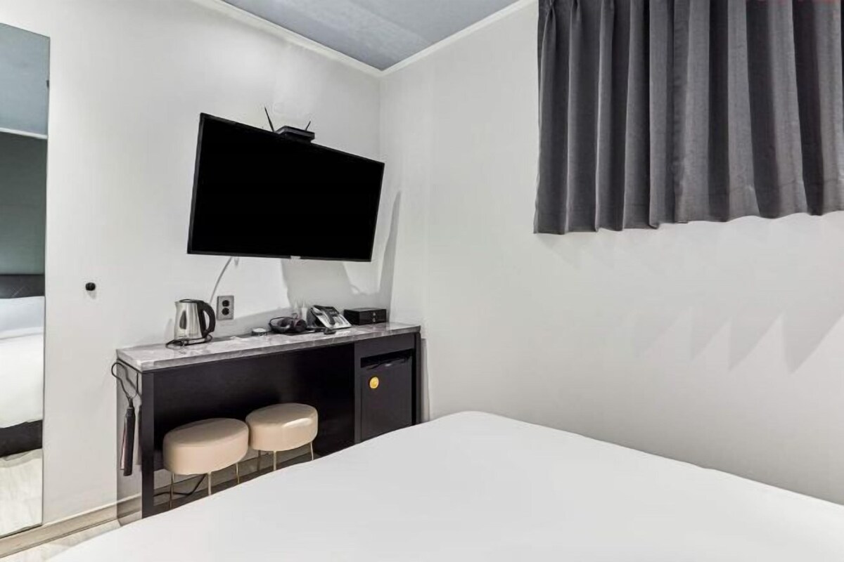 Une chambre d’hôtel comprenant un lit, un bureau avec un téléviseur à écran plat, deux tabourets rembourrés et une fenêtre avec des rideaux.