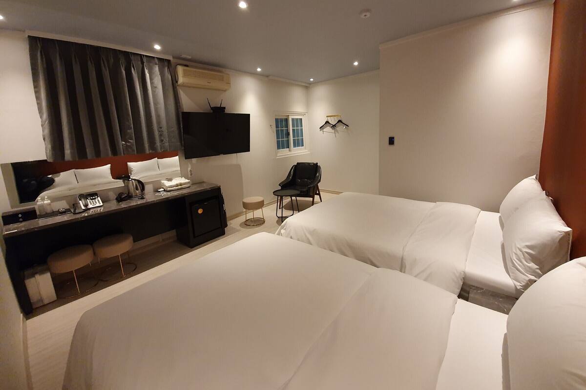 Ein Hotelzimmer mit zwei Betten, einem Schreibtisch mit Telefon und Wasserkocher, einem an der Wand befestigten Fernseher und einem Fenster mit Vorhängen.