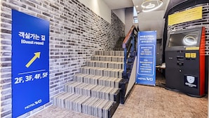 Eine Hotelhalle mit Backsteinwand, einer Treppe zum zweiten Stockwerk und einem blauen Schild, das Zimmer auf den Etagen 2E, 3F, 4F und 5F anzeigt.