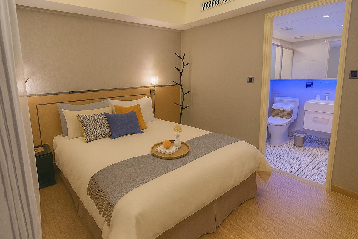 Un lit bien fait, avec un plateau contenant divers objets, une table de chevet et une vue sur une salle de bain équipée de toilettes et d’un lavabo.