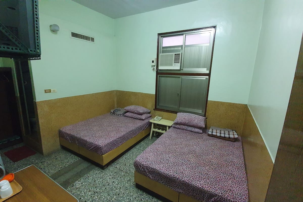 A small, compact room with two single beds, a small table, a telephone, and a window with an air conditioning unit.