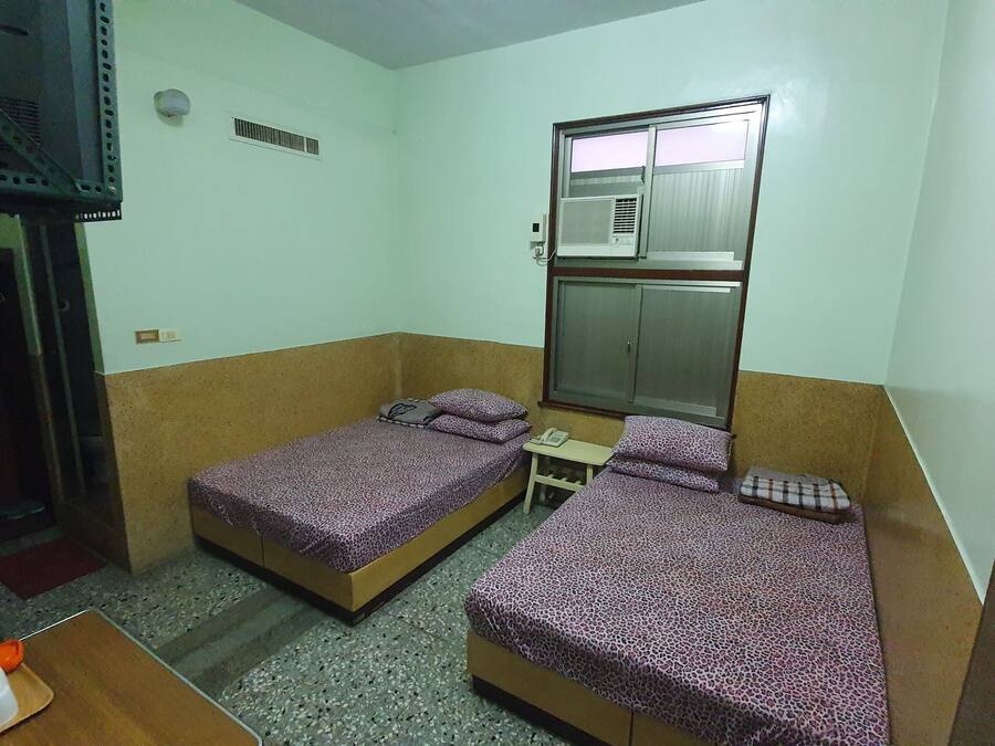 Una habitación pequeña y funcional con dos camas individuales, una mesita, un teléfono y una ventana con unidad de aire acondicionado.