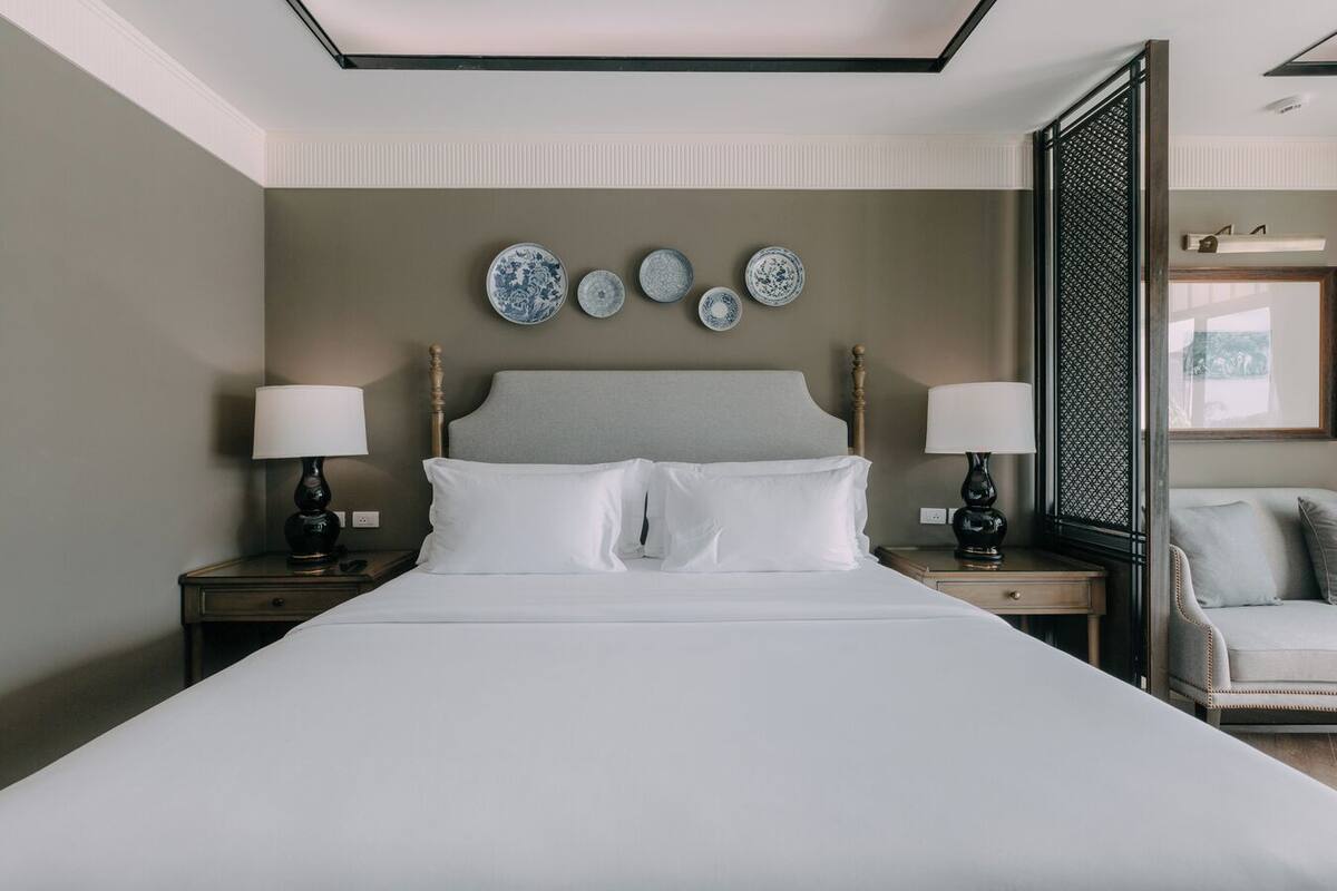 Una cama bien tendida con sábanas blancas, dos mesitas de noche con lámparas, y un arreglo decorativo de platos en la pared.