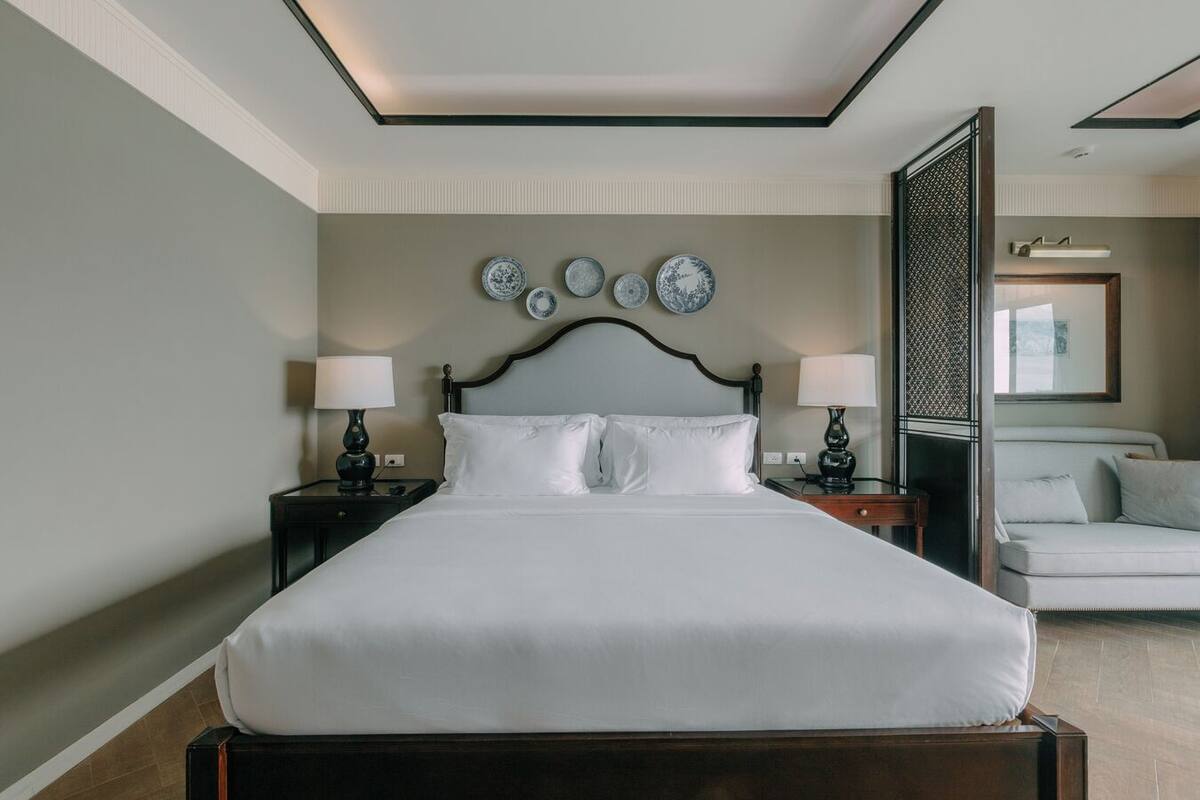 Una cama bien hecha con sábanas blancas, dos mesitas de noche con lámparas y una pared decorada con platos.