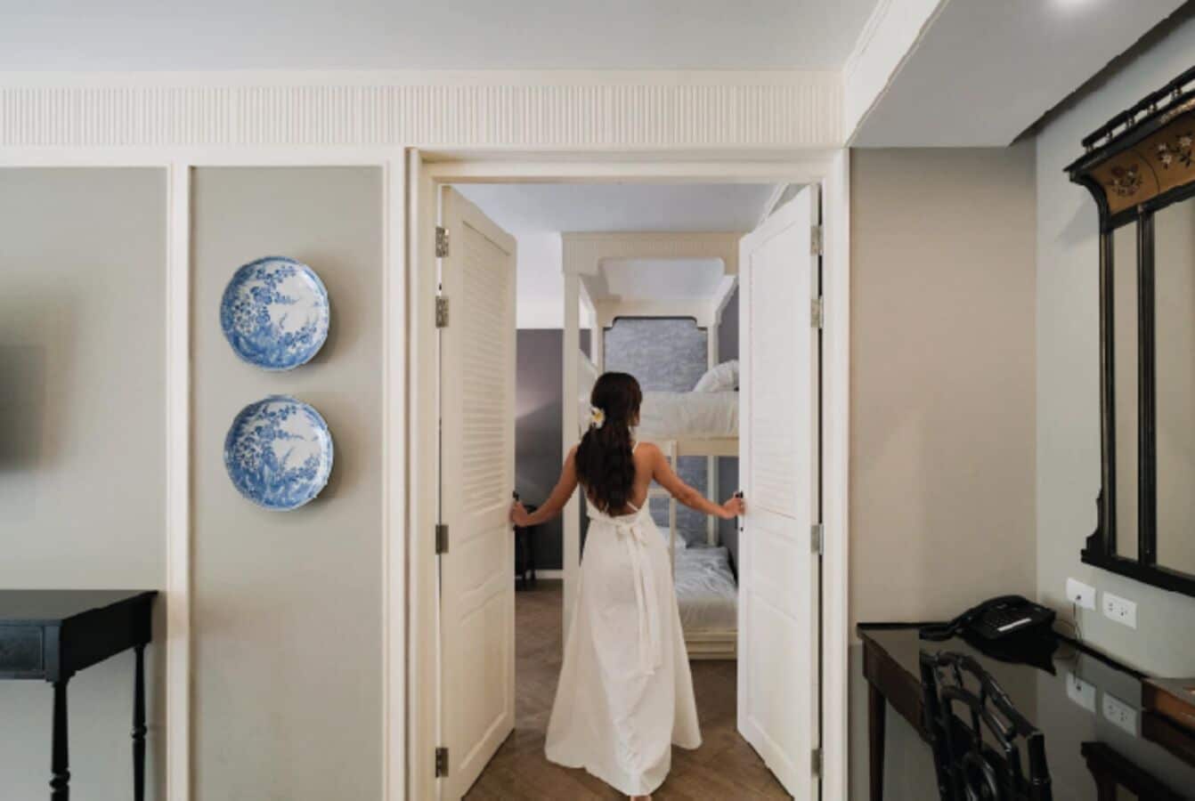 A woman in a white dress walking through a doorway into a bedroom with a bed and a window.