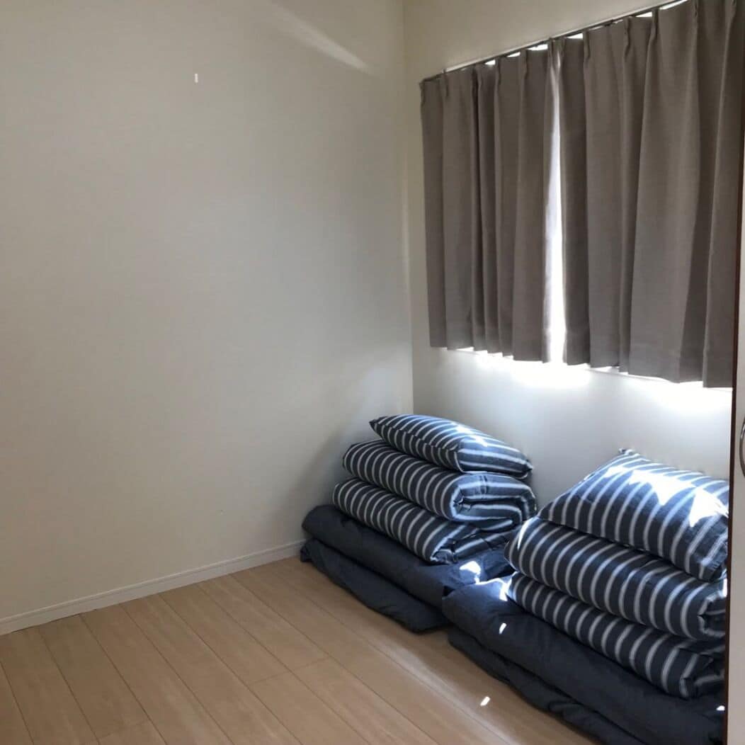 A room with a wooden floor, a window with grey curtains, and a stack of striped pillows.