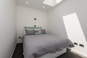 A bedroom with a bed, a nightstand, a wall-mounted heater, and a skylight. - Tree Walk - Redwoods, Spa Pool, Sleeps 10 (Rotorua)