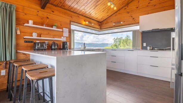 Une cuisine moderne dotée d’un îlot central, de tabourets de bar en bois, de meubles blancs et offrant une vue sur l’extérieur.