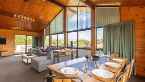 Une pièce spacieuse avec un plafond en bois, de grandes fenêtres, une table à manger dressée pour quatre personnes, un canapé et un téléviseur.