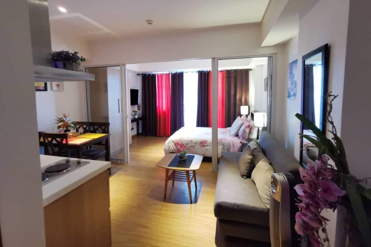 A modern hotel room with a kitchenette, a dining area, a sofa, and a bed with curtains.