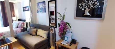 A modern hotel room with a grey sofa, a small wooden table with a vase of flowers, a red trash bin, and a framed artwork on the wall.