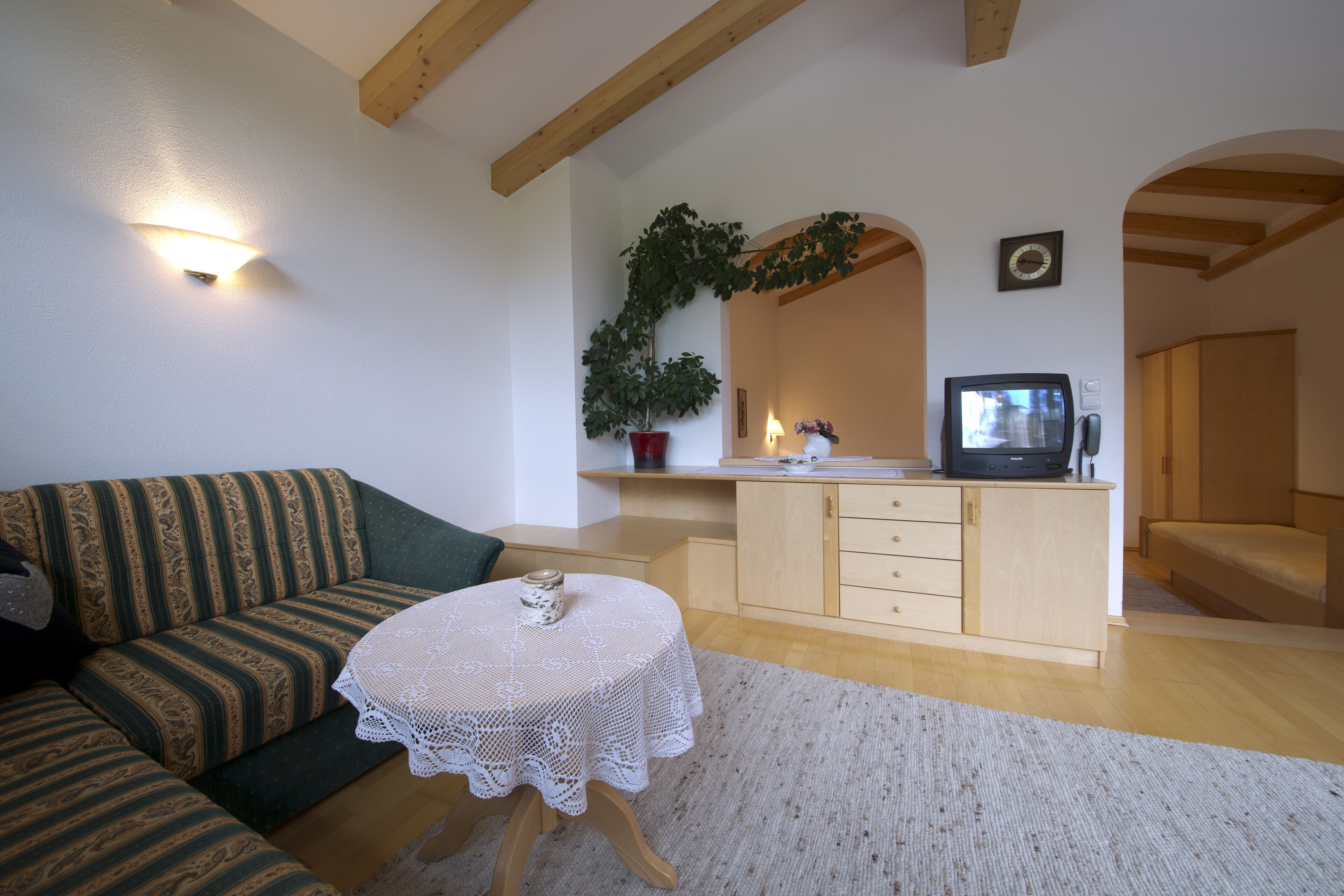 A cozy living room with a green-striped sofa, a small white table with a lace tablecloth, a wooden cabinet, and a potted plant.