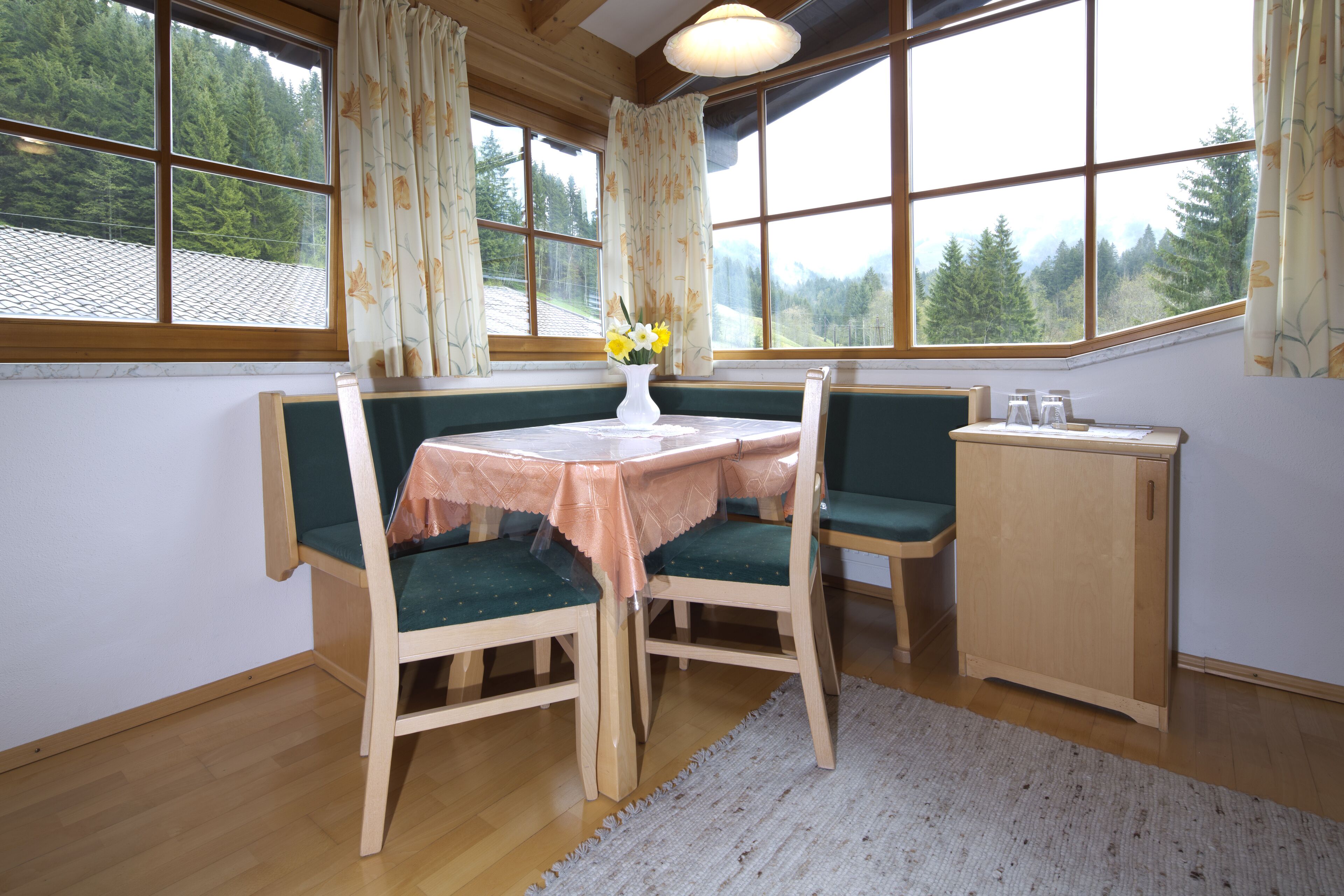 A cozy room with a wooden table and chairs, a small cabinet, and a view of a forest through large windows.