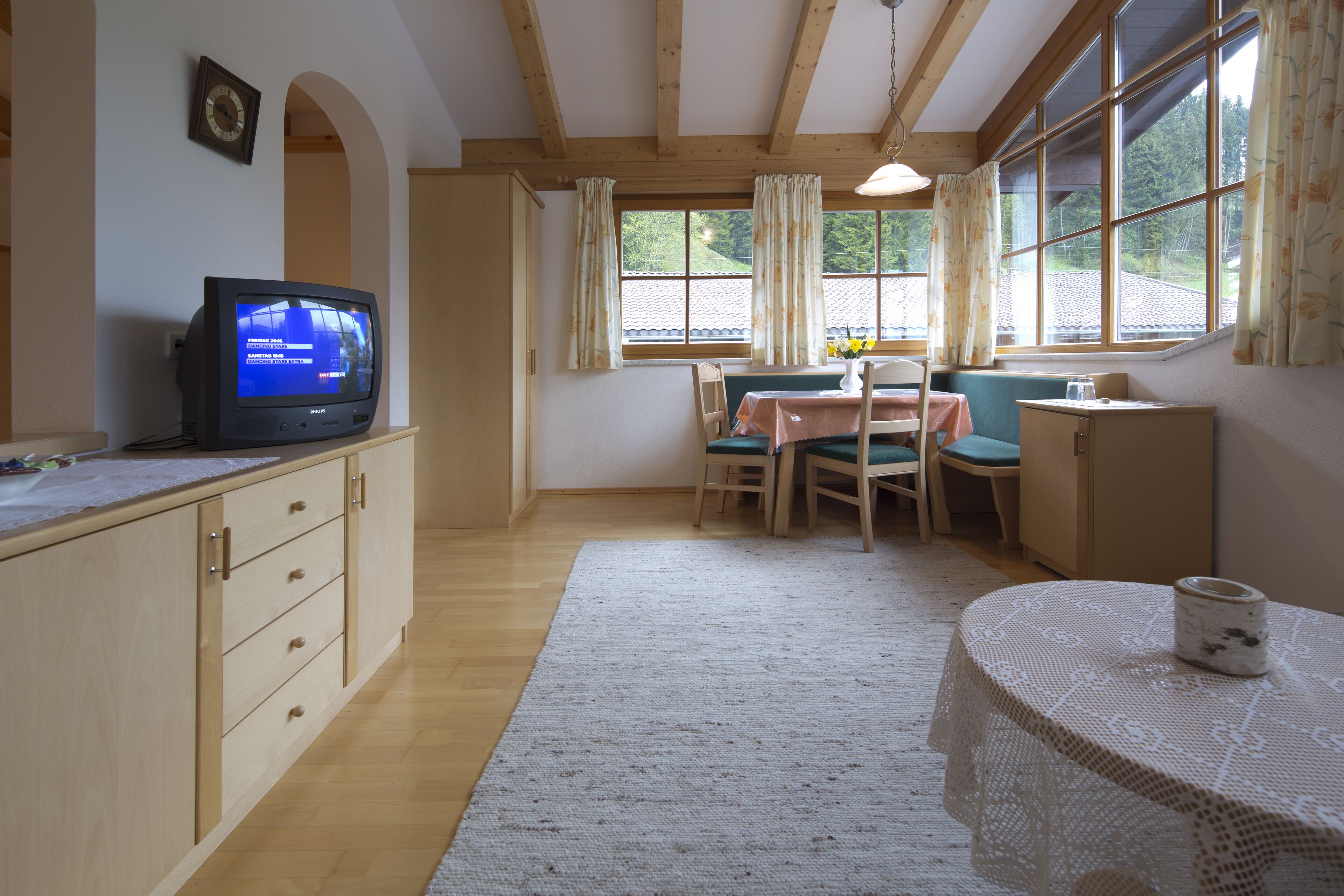 Una habitación acogedora con techo de madera, un televisor sobre un mueble, una mesa de comedor con sillas y una ventana con cortinas.