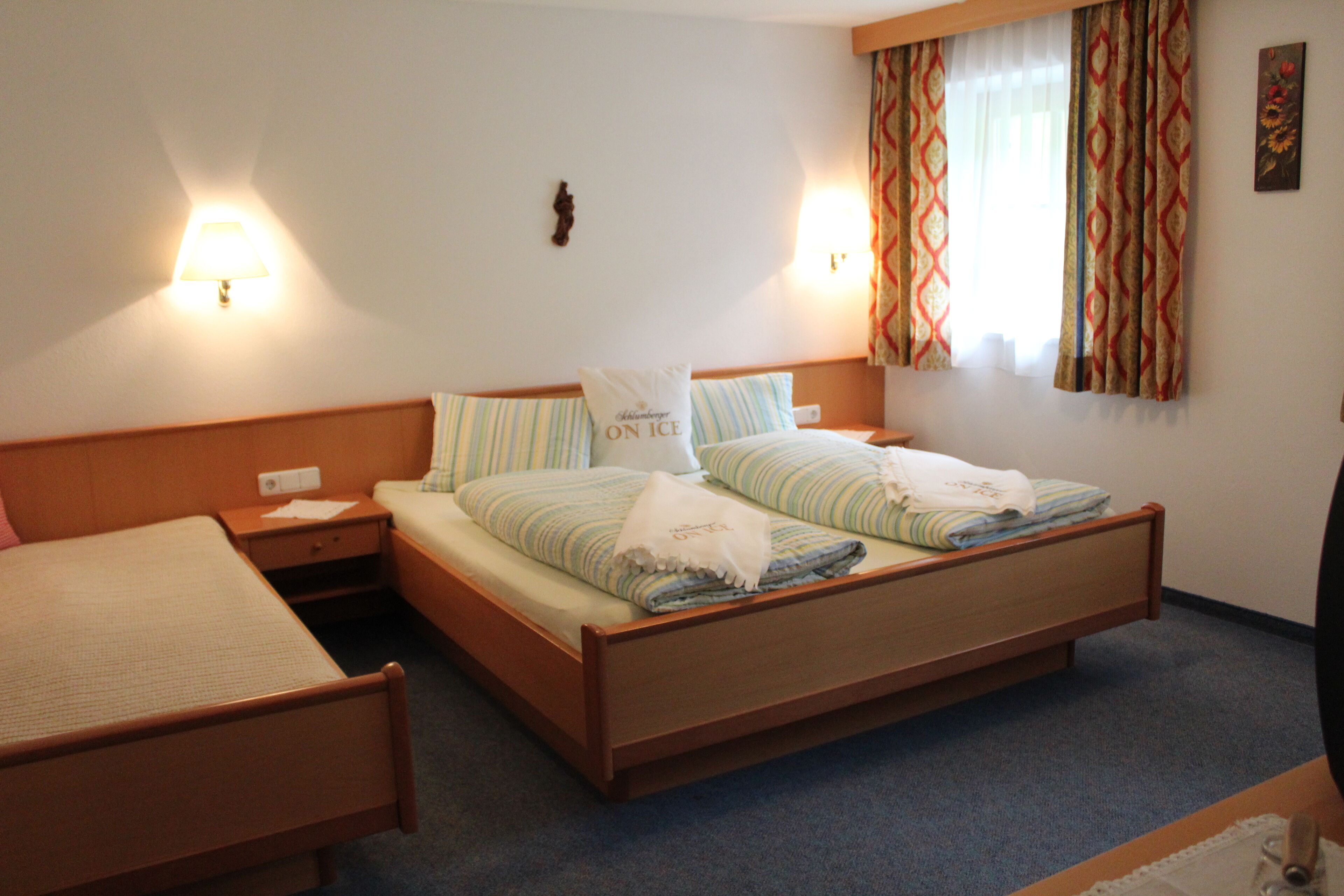 A hotel room with two single beds, a nightstand, a lamp, a window with curtains, and a wall-mounted decoration.