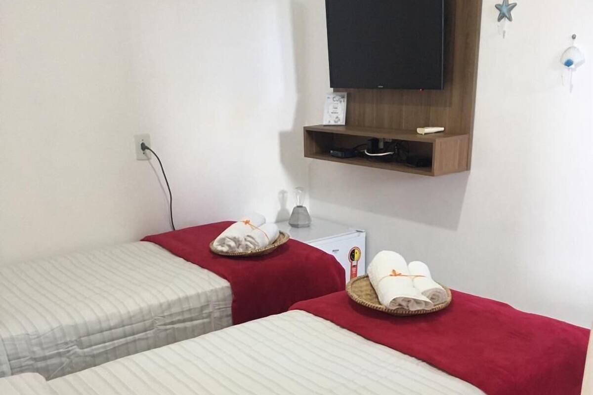 A room with two beds, each with a red blanket and towels on a tray. There is a television mounted on the wall above the beds.