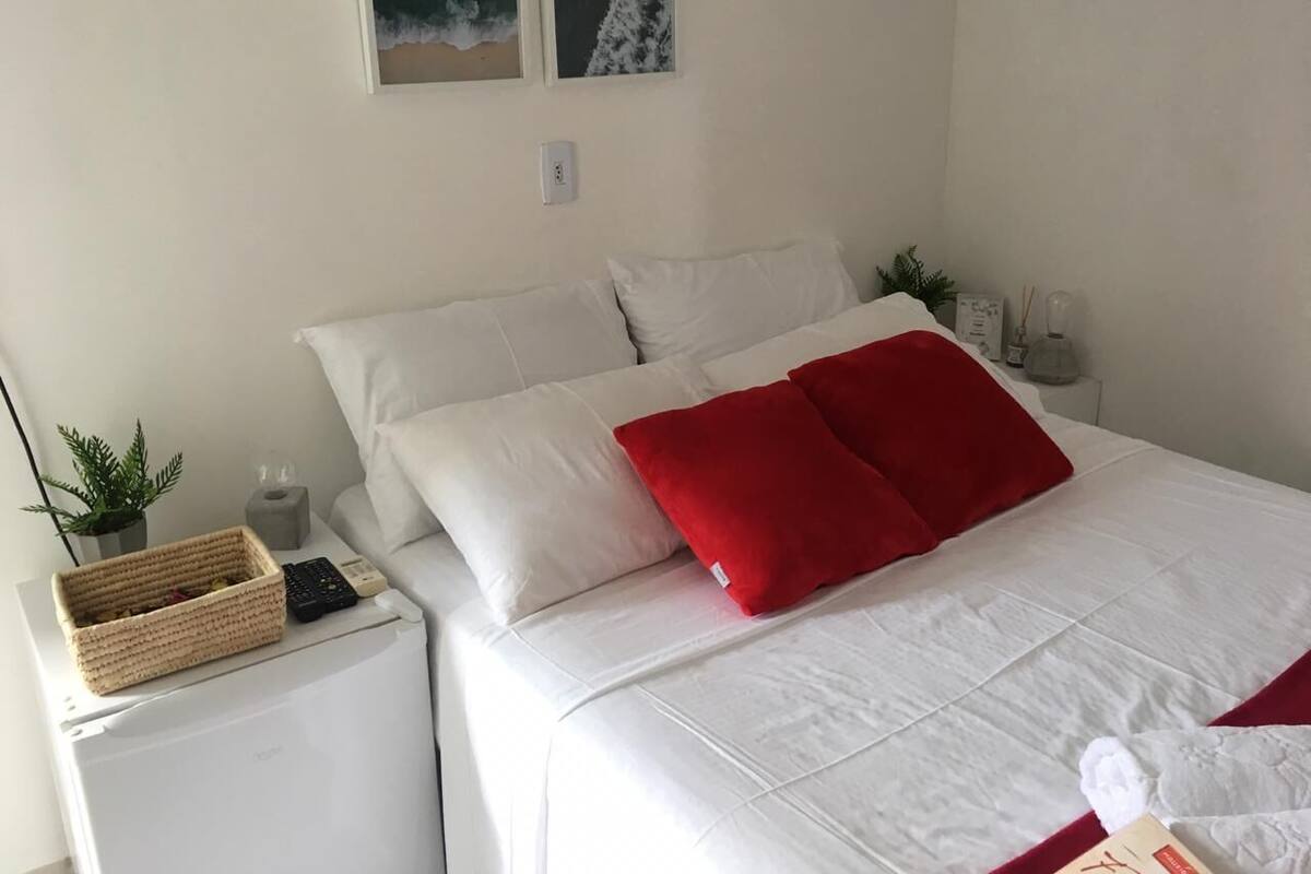 A neatly made bed with white and red pillows, a small bedside table with a remote control, a plant, and a basket.