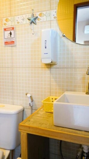 A bathroom with a white sink, a yellow basket, a hand dryer, and a soap dispenser on a tiled wall.