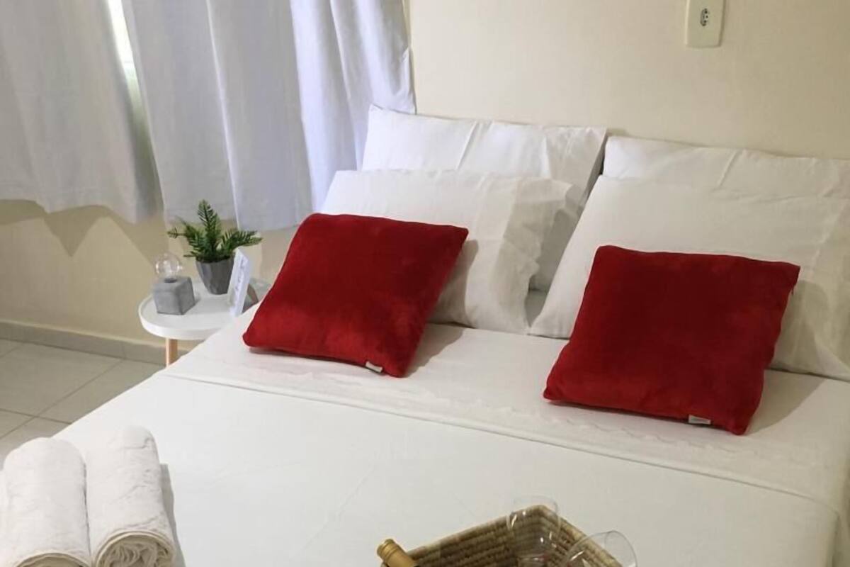 A neatly made bed with white bedding and two red pillows, a small side table with a plant and a bottle, and a basket with a bottle and glasses on the bed.