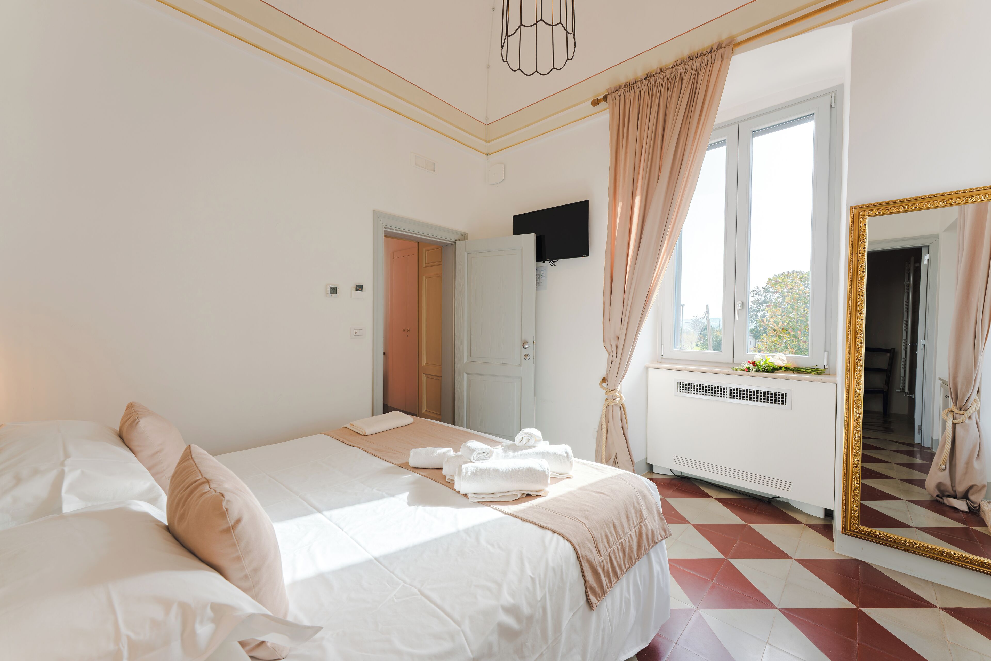 Quarto com uma cama grande, janela com cortinas, radiador e espelho com moldura dourada.