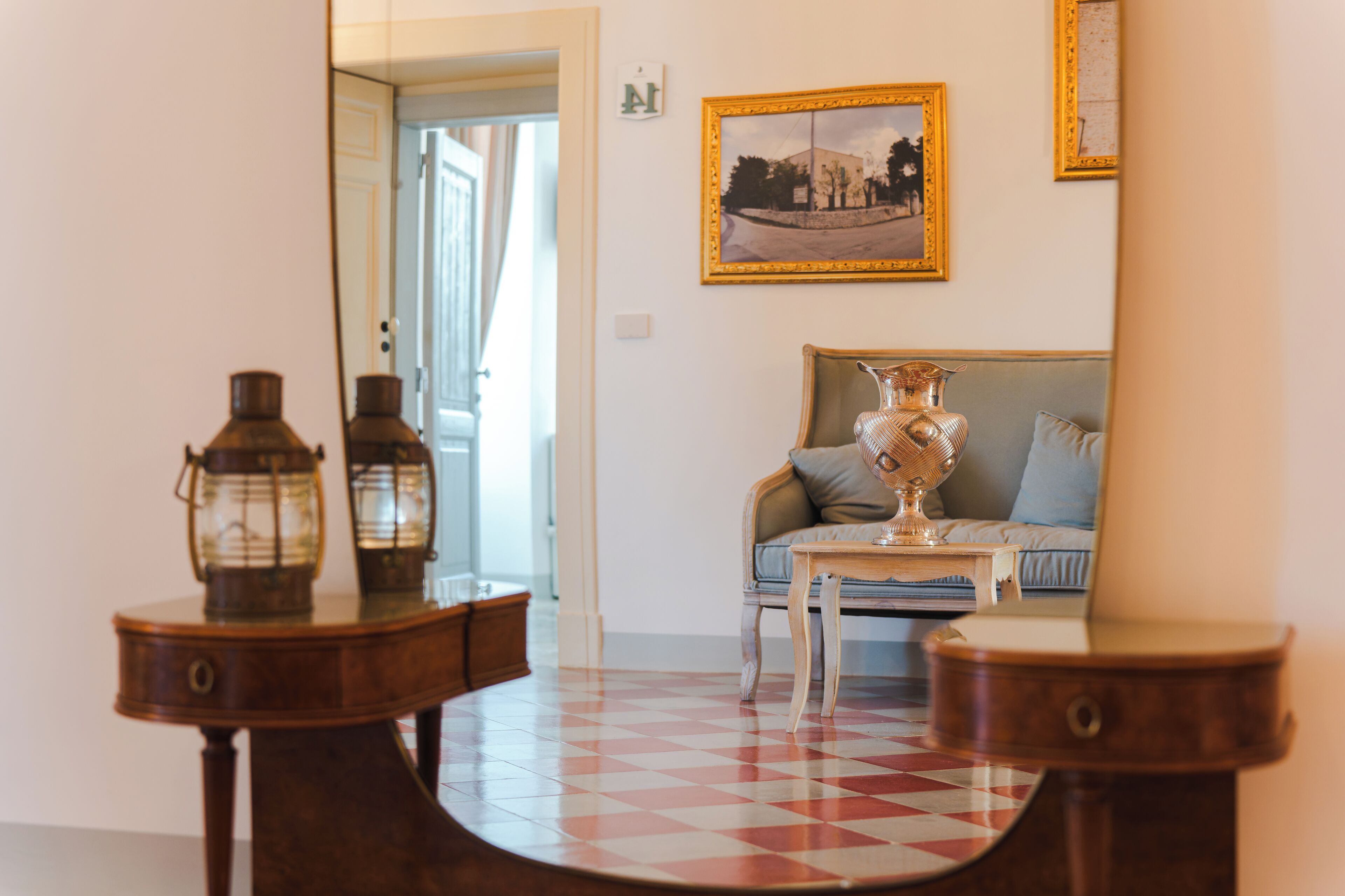 Ein Zimmer mit Schachbrettmuster-Boden, einem Holztisch mit zwei Laternen, einer Couch mit Kissen und einem gerahmten Bild an der Wand.