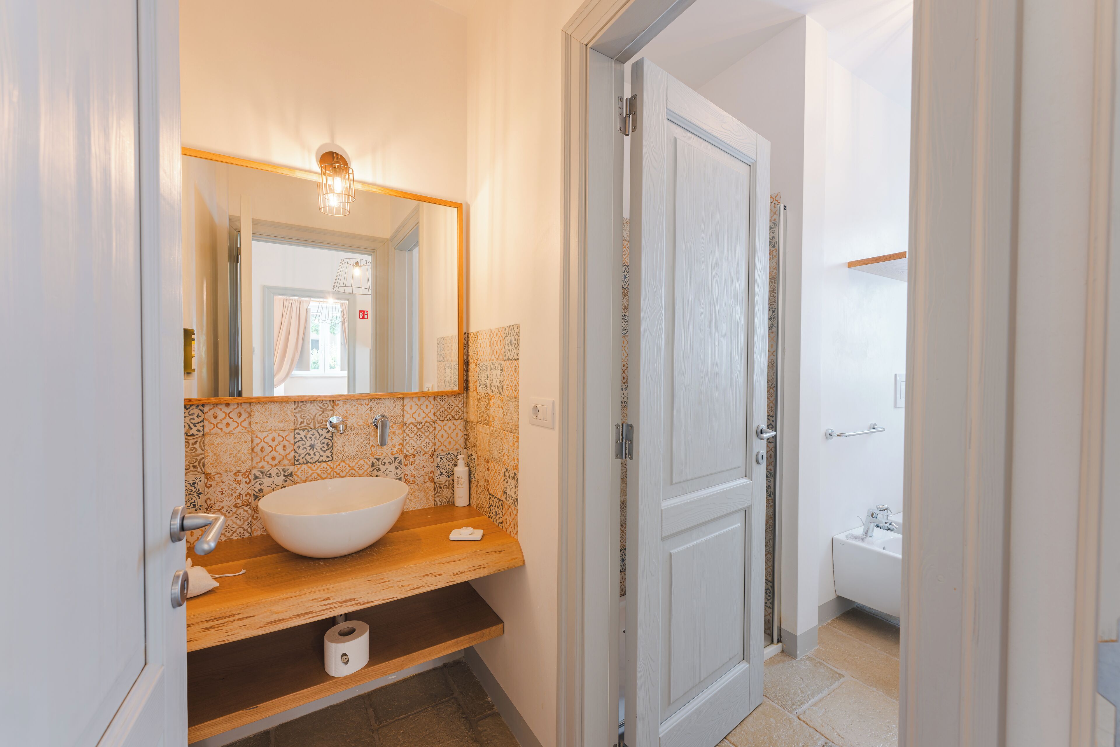 A bathroom with a wooden vanity, a large mirror, a white sink, and a door leading to another room.