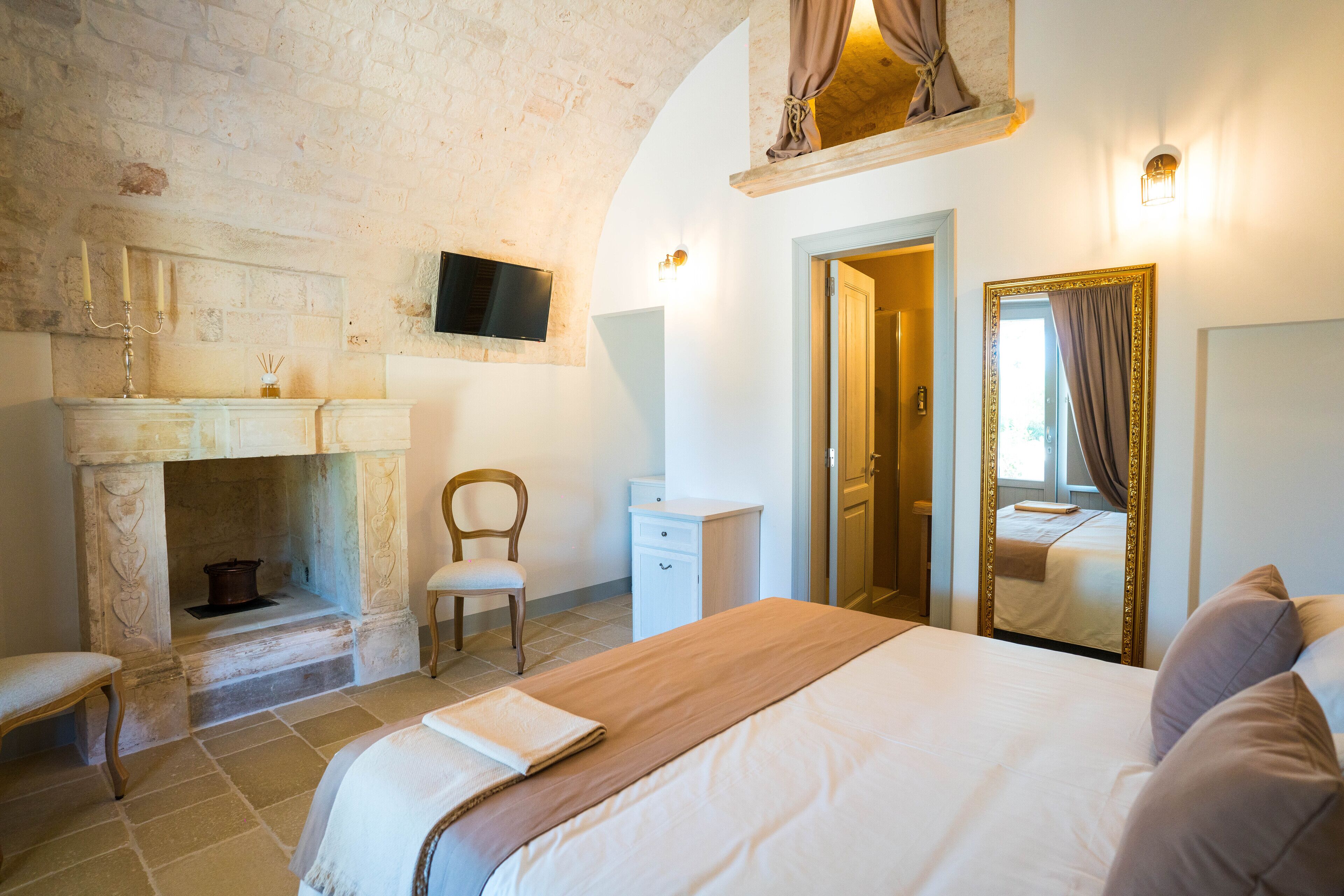 A bedroom with a large bed, a chair, a desk, a mirror, and a fireplace.
