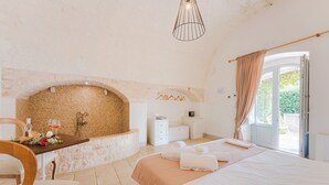 Un dormitorio con chimenea de piedra, una cama con ropa blanca, una silla de madera y una mesita con una botella y vasos.