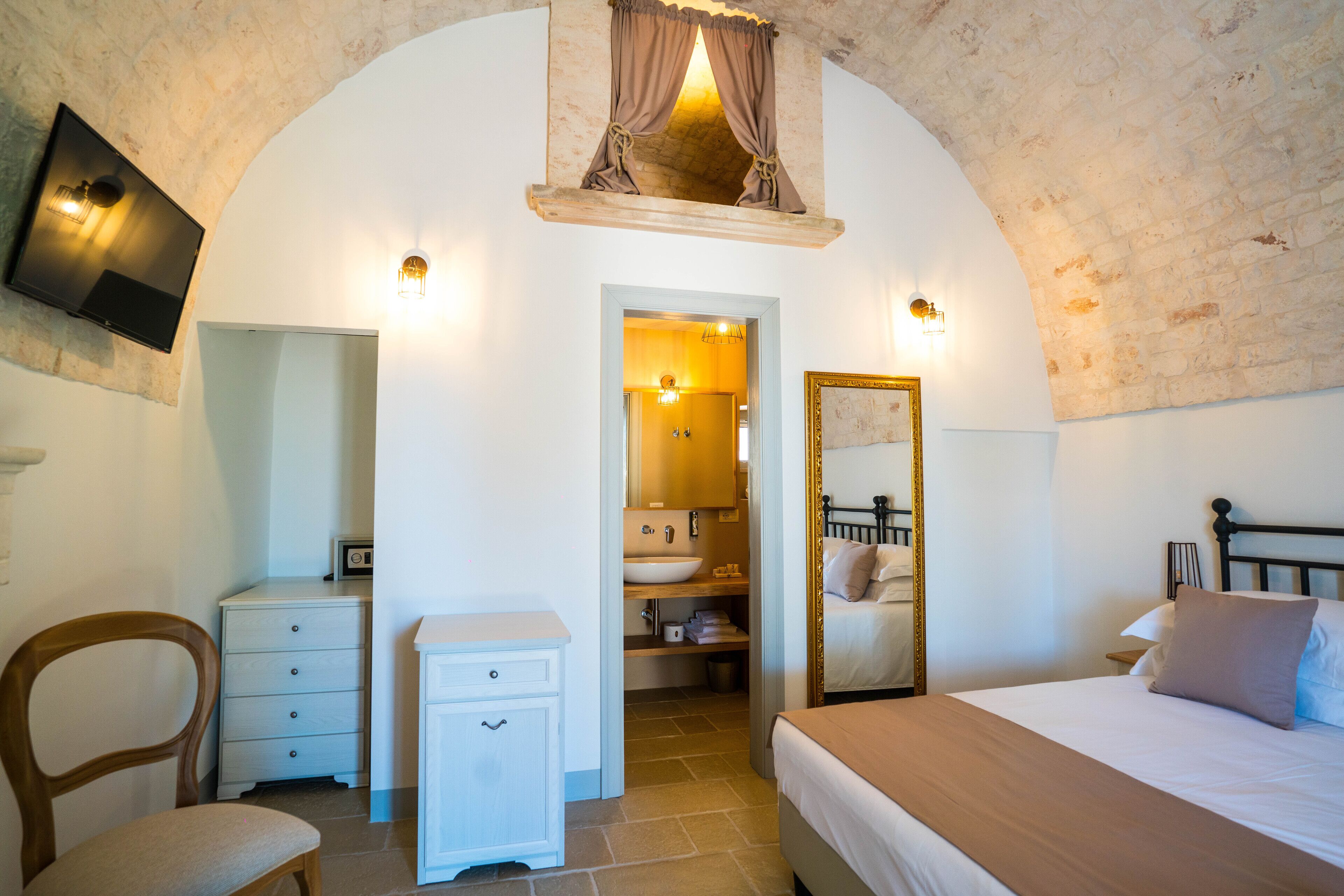 A bedroom with a bed, a nightstand, a chair, a dresser, a mirror, and a bathroom visible through an archway.